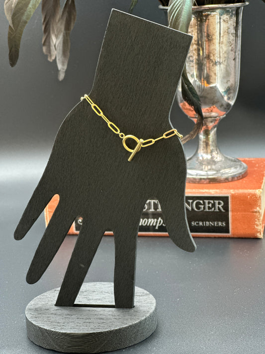 gold paper clip bracelet with toggle chain featured on a black wood model hand with book and silver vase in the background