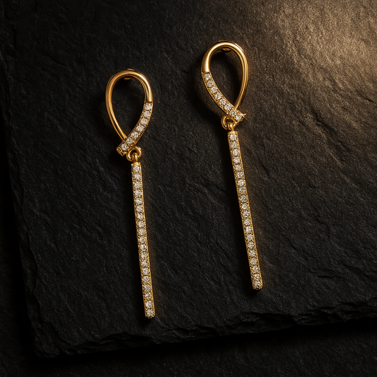 Two matching gold pavé teardrop-bar earrings laid on dark slate granite with warm light from the upper-right corner, highlighting their sparkle.