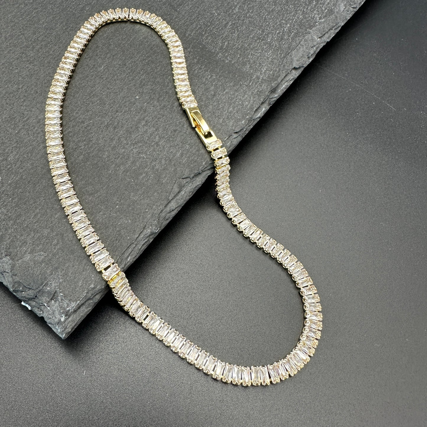 Flat lay of 18K gold-plated baguette tennis necklace showing box clasp positioned toward the front of the neck on a piece of dark gray slate. 