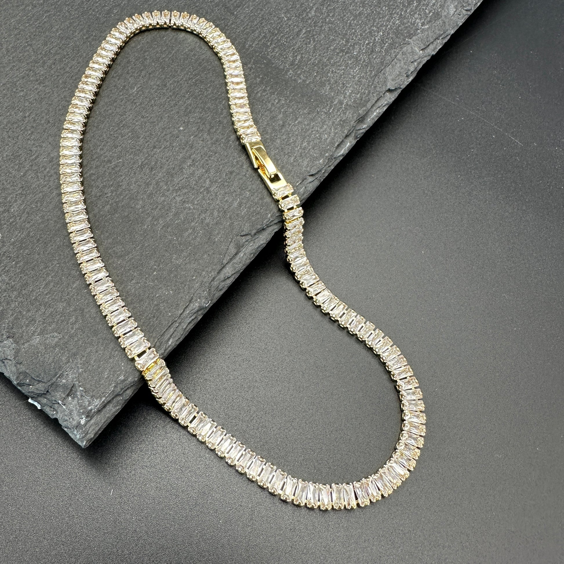 Flat lay of 18K gold-plated baguette tennis necklace showing box clasp positioned toward the front of the neck on a piece of dark gray slate. 